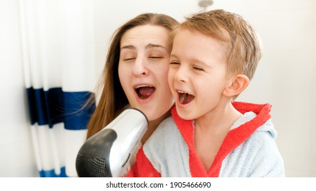 Happy Laughing And Smiling Mother And Little Boy Singing And Having With With Hairdryer In Bathroom. Concept Of Child Hygiene And Health Care At Home. Parents And Kids Having Fun And Playing At Home