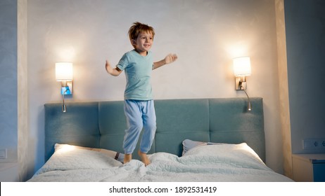 Happy Laughing And Smiling Toddler Boy In Pajamas Skipping And Jumping Up High On Parent's Bed At Night Before Going To Sleep.