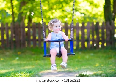 Happy laughing toddler girl with curly hair wearing a blue dress having fun on a swing enjoying a hot sunny summer day on a playground in a park