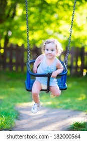 Happy laughing toddler girl with curly hair wearing a blue dress enjoying a swing ride on a sunny summer playground in a park