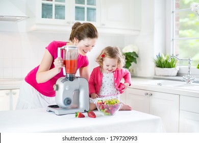 Happy Laughing Toddler Girl And Her Beautiful Young Mother Making Fresh Strawberry And Other Fruit Juice For Breakfast Together In A Sunny White Kitchen With A Window