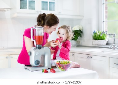 Happy laughing toddler girl and her beautiful young mother making fresh strawberry and other fruit juice for breakfast together in a sunny white kitchen with a window - Powered by Shutterstock