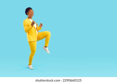 Happy laughing woman dancing cheerfully showing yes gesture clenching her fists isolated on turquoise background, full length. Young stylish african american woman in yellow pants, jacket, sneakers. - Powered by Shutterstock