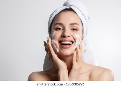 Happy laughing young caucasian woman wrapped in head towel washing face with foam soap cosmetic product. Everyday morning hygiene routine. Skincare and wellness, beauty and cosmetology. Portrait