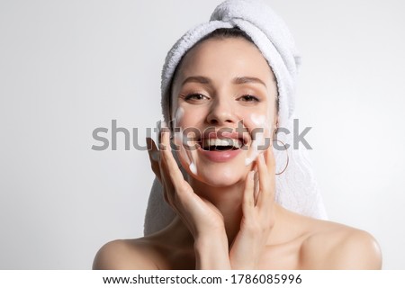 Happy laughing young caucasian woman wrapped in head towel washing face with foam soap cosmetic product. Everyday morning hygiene routine. Skincare and wellness, beauty and cosmetology. Portrait