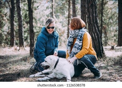 Happy Lesbian Couple Cuddling A Dog While Walking In The Forest