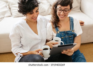 Happy lesbian couple having coffee and using digital tablet in living room. Beautiful women couple at home with a tablet computer. - Powered by Shutterstock