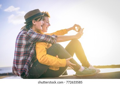 Happy Lesbian Couple Taking A Selfie With Mobile Smart Phone Camera On The Beach At Sunset - Vignette Edit - Homosexuality, Diversity, Vacation, Travel, Lgbt, Technology Concept