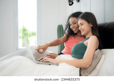 Happy lesbian couple using laptop on bed at home. Young multiethnic lesbian couple using laptop together in bedroom. Technology and love concept - Powered by Shutterstock