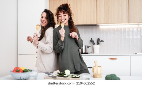 Happy LGBT lesbian couple dancing while cooking together at home - Friends having fun preparing meal in the kitchen - Focus on right girl left hand - Powered by Shutterstock