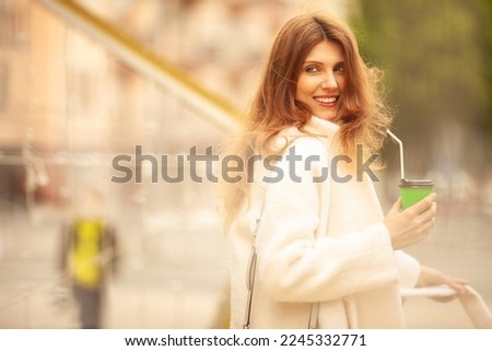 Happy life concept: smiling young woman drinking hot beverage through sipper at street of Mariupol city, Ukraine. Wind blown long hair. Model wearing white coat. Copy-space. Outdoor shot