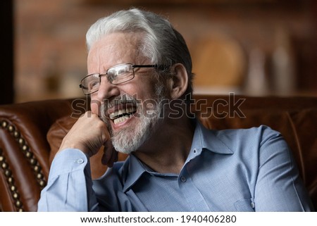 Happy life on pension. Mature aged man retiree wear glasses sit on sofa look aside laugh demonstrate healthy straight teeth prothesis implants. Old man with white smile advertise dental clinic service