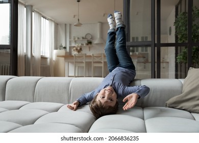 Happy little 7s kid boy lying on comfortable sofa upside down, having fun entertaining alone in modern living room. Smiling adorable small child enjoying leisure active pastime, playing at home.