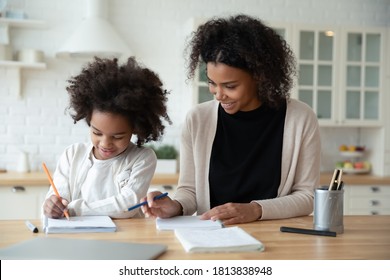 Happy little adorable african american girl enjoying studying with mommy's assistance at home. Smiling caring mixed race woman helping small school aged child daughter preparing homework indoors.