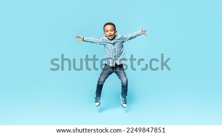 Happy little african american boy posing in mid air, jumping and spreading arms over blue studio background, full length. Carefree child having fun. Kids fashion and style