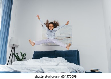 Happy Little African American Girl In Pajamas Jumping On Bed