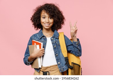 Happy little african american kid school girl 12-13 years old in casual denim clothes backpack hold books show victory v-sign gesture isolated on pastel pink background Childhood education concept