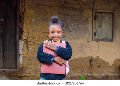 A happy little African girl child or student with pink school bag, holding and hugging her books outside a village mud house