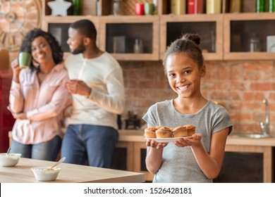 Happy little african girl demonstrating fresh home made cupcakes, mommy and daddy talking on background, kitchen interior, empty space - Powered by Shutterstock