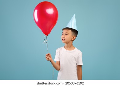 Happy Little Asian B-Day Boy Wearing Party Hat Holding Red Balloon And Looking On It, Excited Small Male Child Celebrating Birthday, Standing Isolated Over Blue Studio Background, Copy Space