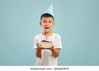 Happy Little Asian Boy In Party Hat Holding Piece Of Birthday Cake With Candle, Excited Cheerful Male Kid Celebrating B-Day, Making Wish While Standing Over Blue Studio Background, Copy Space