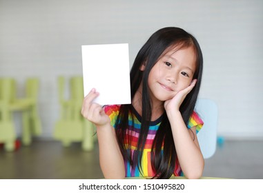 Happy Little Asian Child Girl Holding Blank White Paper Card In Her Hand. Kid Showing Empty Paper Note Copy Space In Children Room.