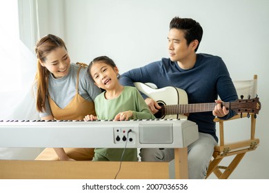 Happy Little Asian Family Daughter Playing Piano With Mother And Father Play Guitar At Home, Mother Teaching Daughter To Play Piano,They Play And Sing Songs. Concept For Family Relationship.