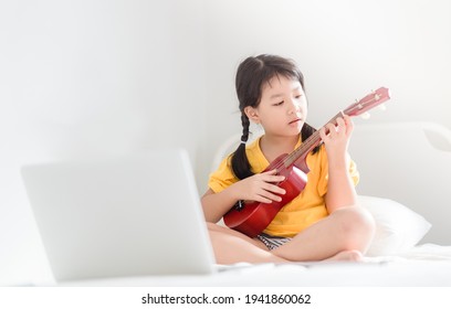 Happy Little Asian Girl Sing A Song Playing Ukulele Online Learning Music Ukulele Guitar Class At Home.Online Education, Music Art.Homeschool Kid Asian Child Girl Live Streaming Laptop In Bedroom.