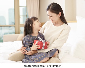 Happy Little Asian Girl Sitting In Arms Of Young Loving Mother Holding  A Wrapped Box Of Gift