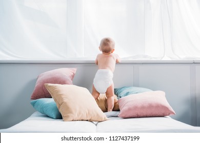 happy little baby in diaper standing on bed with lot of pillows and looking through window