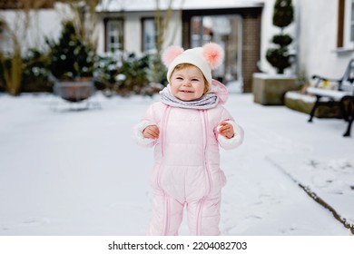 Happy little baby girl making first steps outdoors in winter through snow. Cute toddler learning walking. Child having fun on cold snowy day. Wearing warm baby pink clothes snowsuit and bobbles hat.