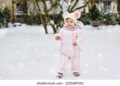 Happy little baby girl making first steps outdoors in winter through snow. Cute toddler learning walking. Child having fun on cold snowy day. Wearing warm baby pink clothes snowsuit and bobbles hat