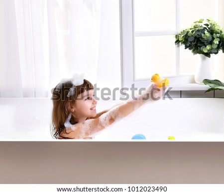  Happy little baby girl sitting in bath tub  in the bathroom with little ducks. Portrait of baby bathing in a bath full of foam near window