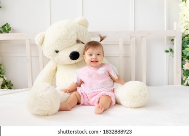 happy little baby girl six months old sitting on a white bed in pink clothes, with a big Teddy bear and smiling