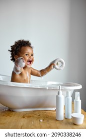 Happy Little Black Baby Girl Sitting In Bath Tub Playing With Toys In Bathroom. Portrait Of Baby Bathing In White Bath Full Of Foam, At Home. Everyday Life. Hygiene, Children, Lifestyle Concept