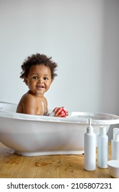 Happy little black baby girl sitting in bath tub playing with toys in bathroom. Portrait of baby bathing in white bath full of foam, at home. everyday life. hygiene, children, lifestyle concept