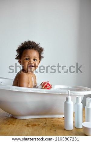 Happy little black baby girl sitting in bath tub playing with toys in bathroom. Portrait of baby bathing in white bath full of foam, at home. everyday life. hygiene, children, lifestyle concept