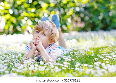 Happy little blond child with blue eyes laying on the grass with daisies flowers in the park. On warm summer day. Kid boy dreaming and smiling.