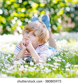 Happy little blond child with blue eyes laying on the grass with daisies flowers in the park. On warm summer day. Kid boy dreaming and smiling.