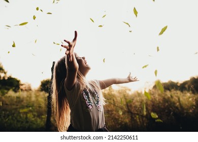 Happy Little Blonde Girl Plays With Green Leaves In Garden, Field. The Child Smiles, Laughing And Has Fun In Park. Playful Kid Throwing Up Leaves In Summer Or Autumn Season. Child Lays In Country Side