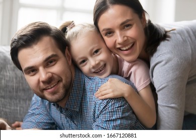 Happy little blonde kid girl lying on fathers back with attractive young mother, head shot close up portrait. Beautiful couple posing for family photo with cute little child daughter, resting on sofa.