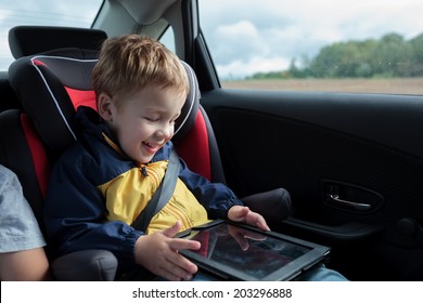 Happy little boy in the car sitting in child safety seat and playing with tablet PC