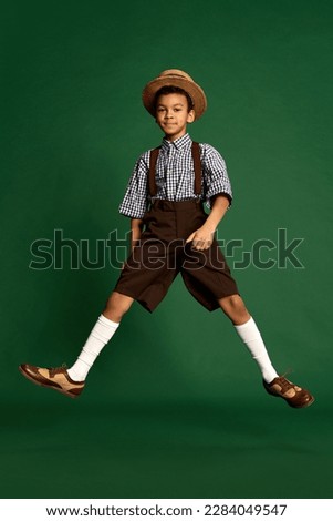 Happy little boy, child in checkered shirt and suspender shorts jumping, having fun over dark green background. Concept of happiness, childhood, friendship, retro fashion style