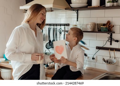 Happy little boy congratulating smiling mother and giving card with red heart during holiday celebration at home. Family life events celebration, Mother Day congrats concept