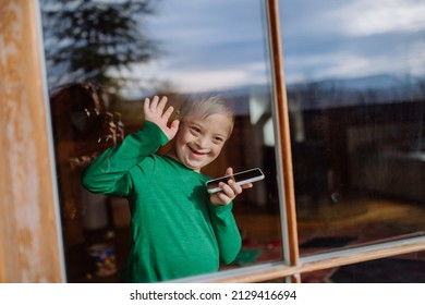 Happy Little Boy With Down Syndrome Using Smartphone And Waving Through Window