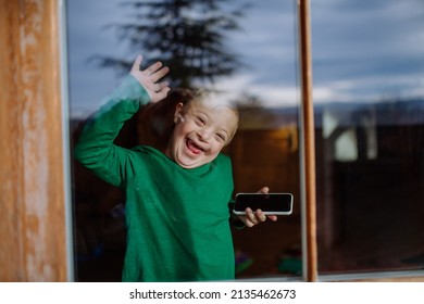 Happy Little Boy With Down Syndrome Using Smartphone And Waving Through Window