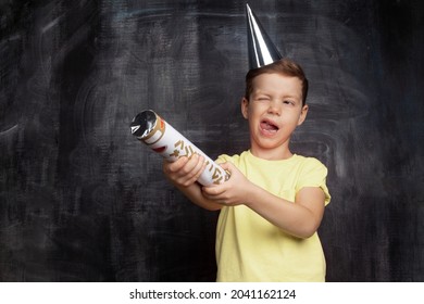 Happy little boy with flapper celebrating holiday, popping cracker during birthday party. Playful child in festive cap with slapstick shooting popper on background of black chalk board with copy space