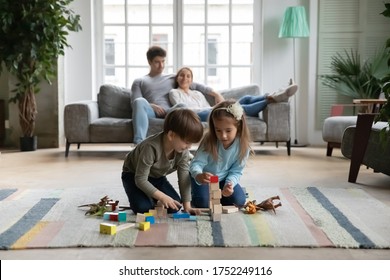 Happy little boy and girl kids sit on warm floor in living room building with wooden colorful blocks bricks, young parents relax on comfortable sofa in background, Caucasian family leisure weekend