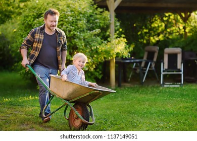 Happy little boy having fun in a wheelbarrow pushing by dad in domestic garden on warm sunny day. Active outdoors games for kids in summer. - Powered by Shutterstock