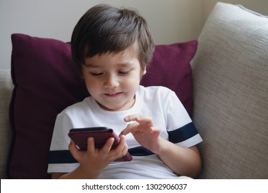 Happy Little Boy Having Fun Playing Game On Mobile Phone,Preschool Kid Sititng On Sofa With Smiling Face Watching Cartoon On Smart Phone,Child Using Cell Phone While Relaxing At Home